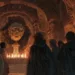 La tête momifiée d'Henri IV repose sur un piédestal au centre d'une crypte gothique sombre, éclairée par des bougies. Autour, des figures en capes noires observent avec une intense curiosité. L'architecture complexe et les sculptures anciennes ajoutent à l'atmosphère mystérieuse de la scène.