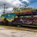 Un corbillard classique paré de peintures murales colorées célébrant le jazz est garé devant un grand graffiti mural dans un style similaire, illustrant le riche héritage culturel et artistique du corbillard dans la société contemporaine.