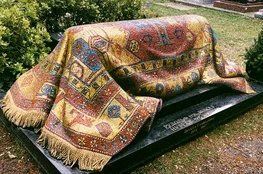 Sépulture de Rudolf Noureev recouverte d’un kilim oriental en mosaïque, photographiée dans un cimetière arboré. Le tombeau, en pierre noire, est orné de motifs colorés rappelant les tapis d’Orient.